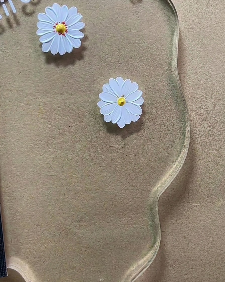 A fine detailing brush applying tiny reddish-brown dots to the yellow center of a white painted daisy on a clear acrylic palette.