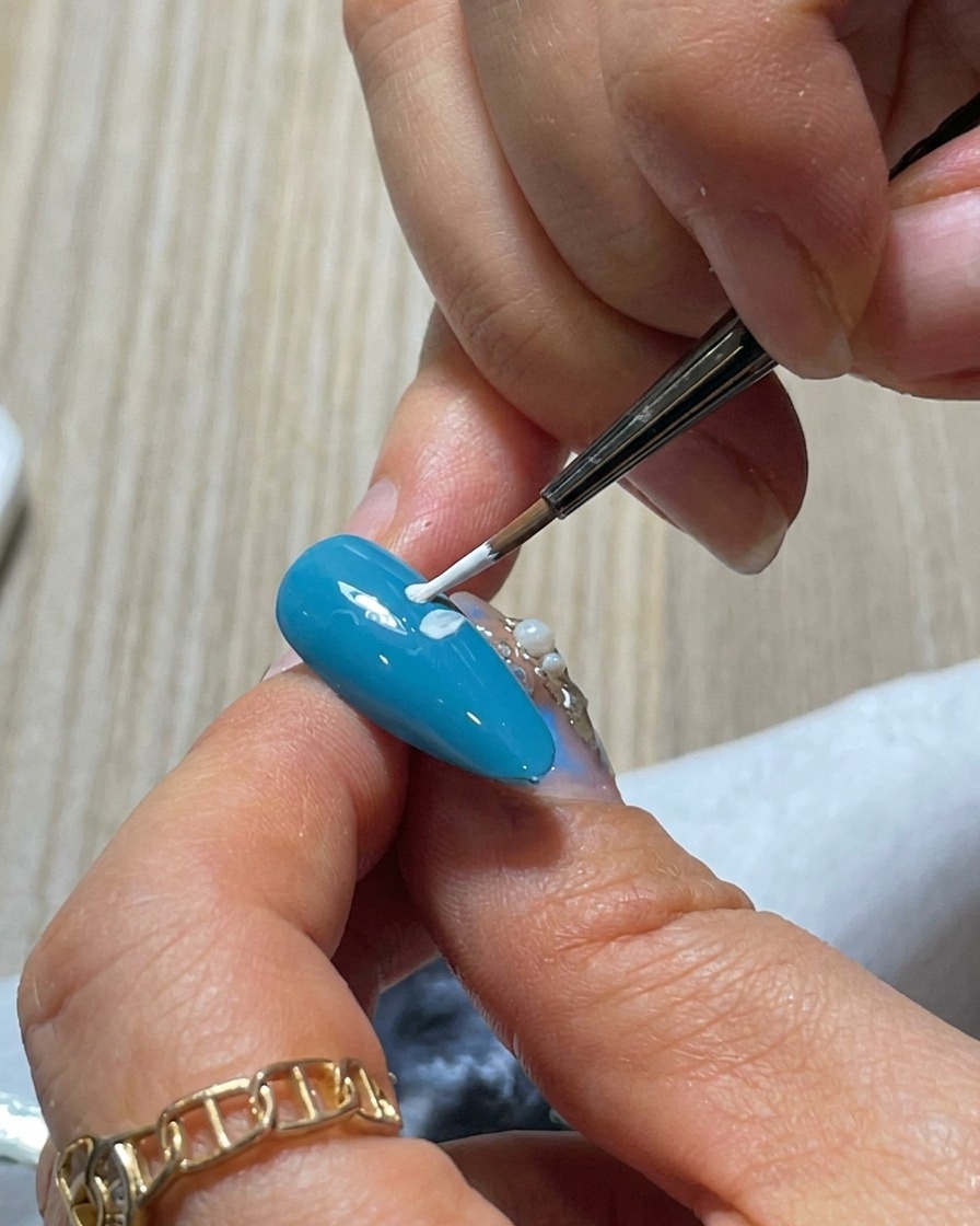 A close-up view showing a third white petal being added to the blue nail, completing part of the daisys outer ring.