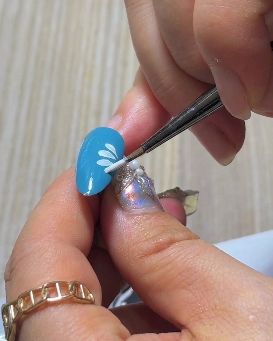 A close-up view of a brush painting the final white petal to complete the bottom layer of a small daisy design on a blue nail.