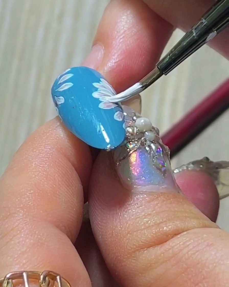 A close-up of a brush adding a third white petal to the overlapping daisy layer on a blue nail.