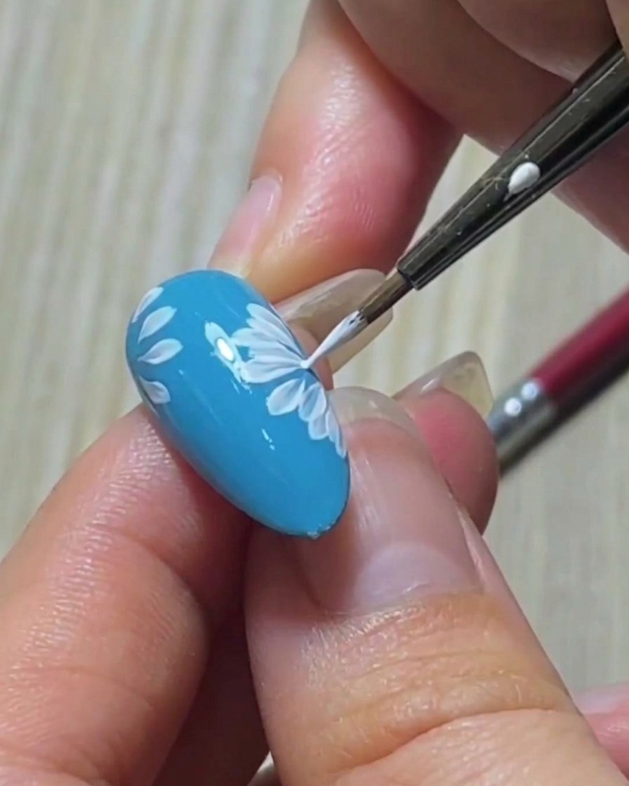 Close-up of a liner brush refining the sharp point of a white petal on a blue nail base.