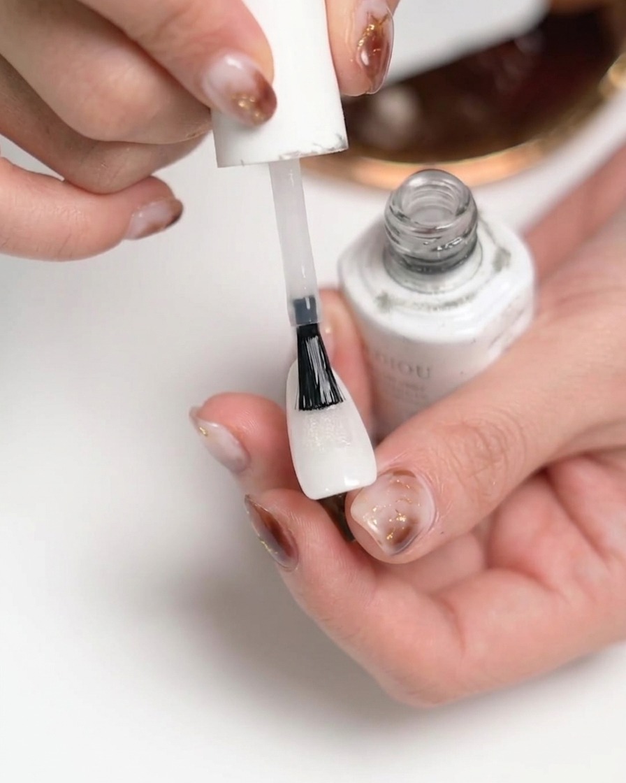 A close-up of a hand applying a clear matte top coat to a white-painted fingernail using a brush from a white polish bottle.