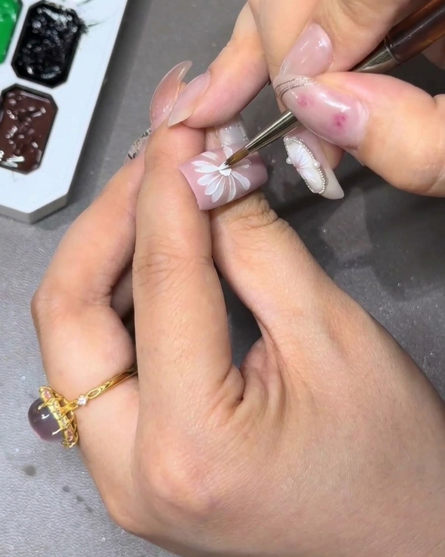 A close-up of a nail being decorated with a complete, multi-layered white flower design.