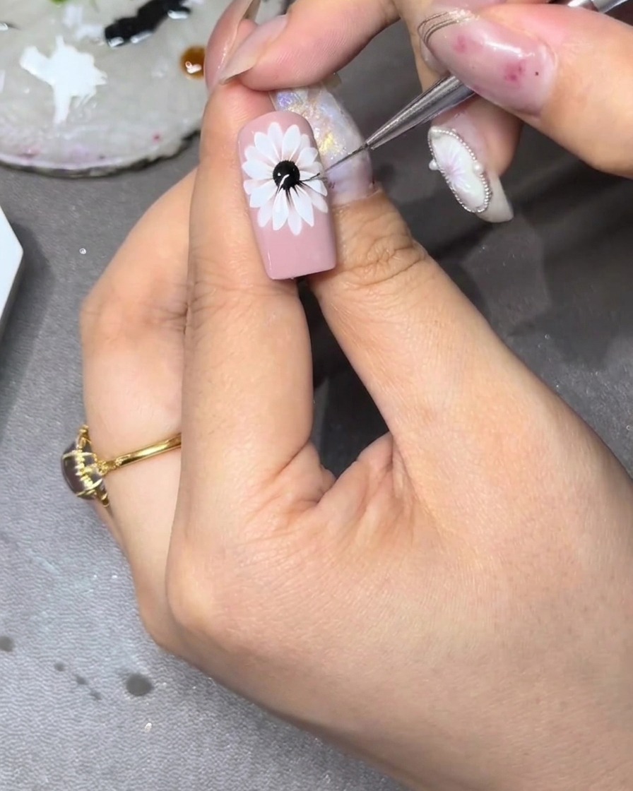 A close-up of a nail with a white floral design, where a fine brush is flicking small yellow strokes outward from the center into the spaces between the white petals.