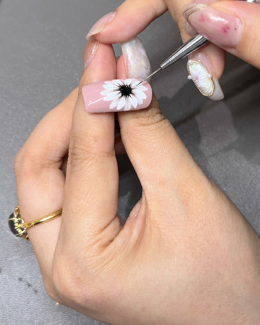 A fine detail brush refining the dark black center of a painted white daisy on a light pink nail.