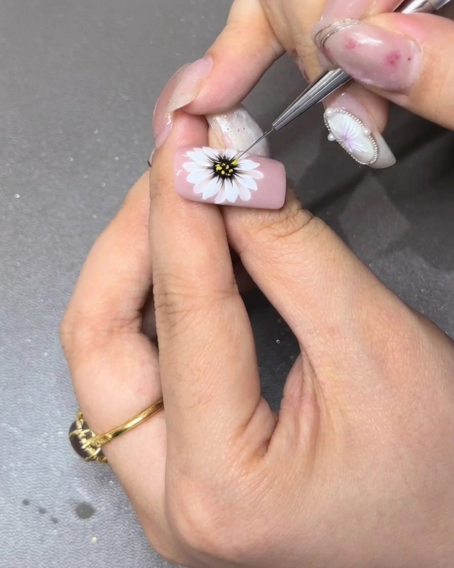 A fine liner brush adding tiny yellow stamen dots around the black center of a painted daisy.