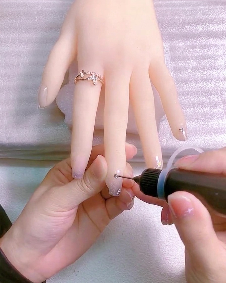 A fine brush applying clear gel around the base of a crystal charm on a fingernail to secure it.