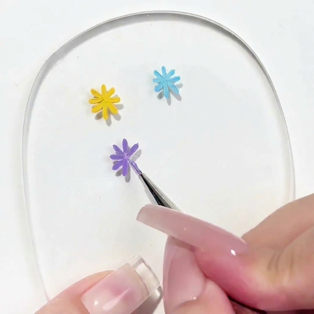 A close-up view of a detail brush applying a petal to a partially completed purple daisy on a clear nail art palette.