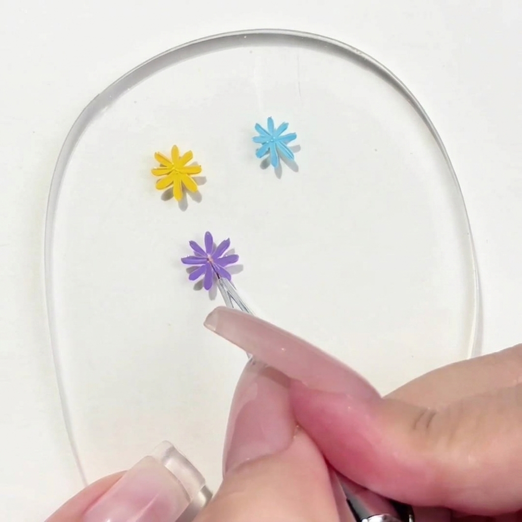 A close-up of a detail brush placing a small, yellow dot in the center of a hand-painted purple daisy.