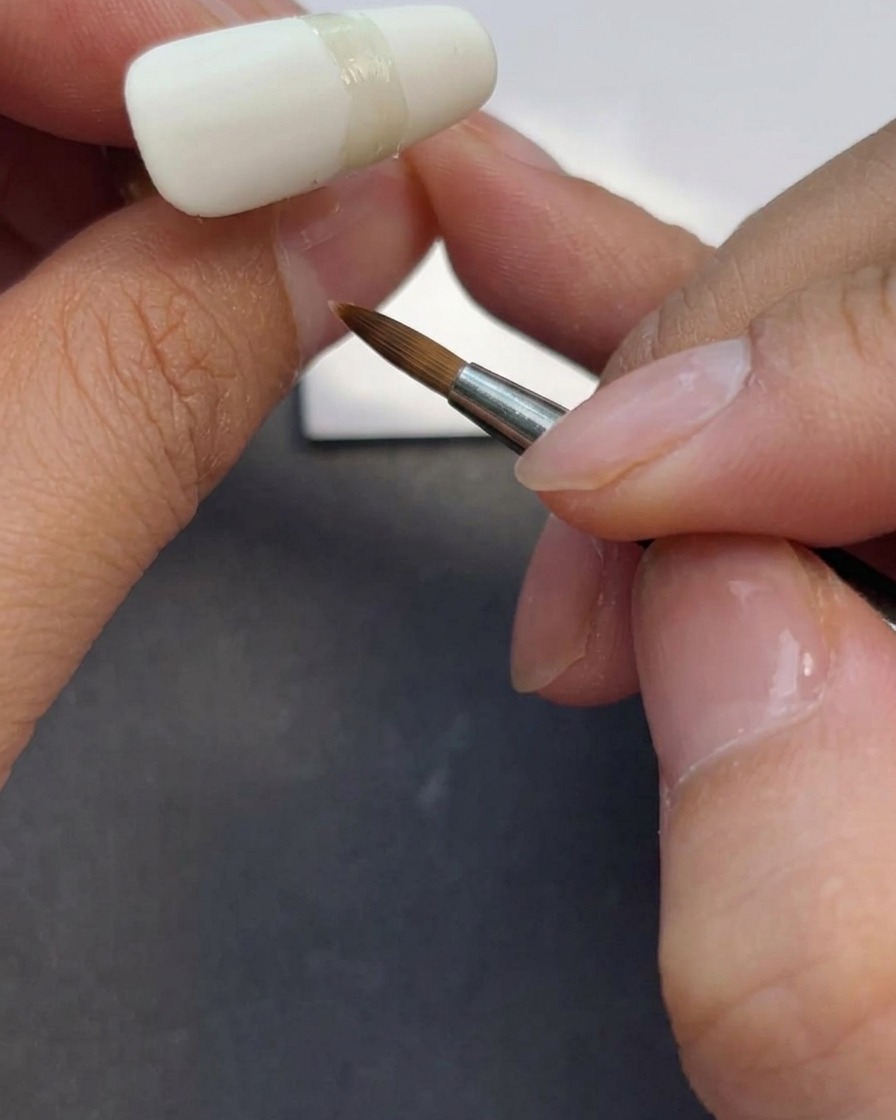 A nail technician using a gel brush to apply a layer of clear matte top coat over a milky white fingernail.