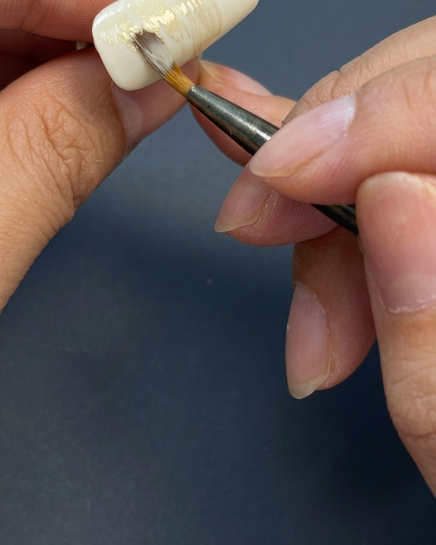 A brush applying light, streaky horizontal strokes of mixed earthy colored gel onto a milky white nail to create a textured background.