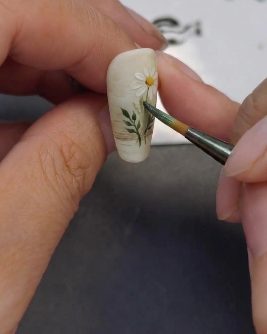Close-up showing a fine brush adding small green leaves to the painted flower stems on the nail.