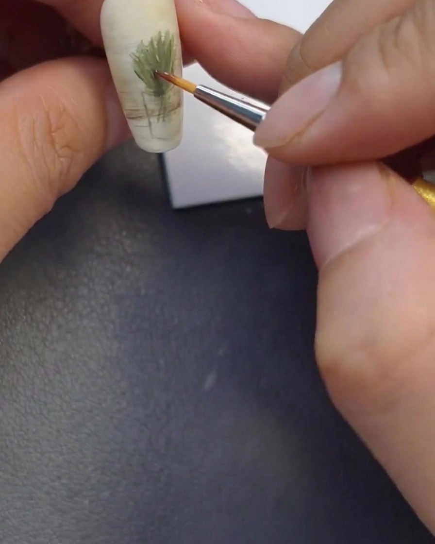 Close-up of a fine brush applying small dots of brown, orange, and yellow to the nail to form flower centers.