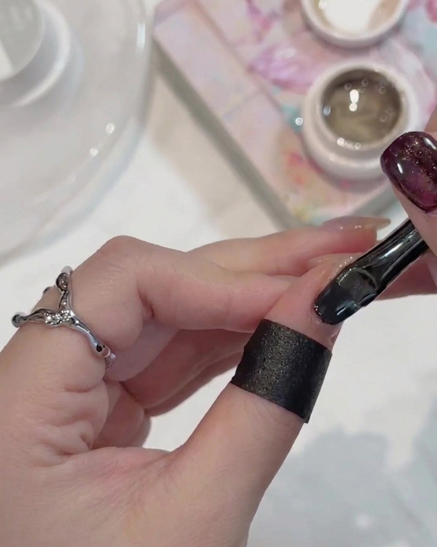 A nail brush applying dark, shimmering cat-eye gel polish to a fingernail.