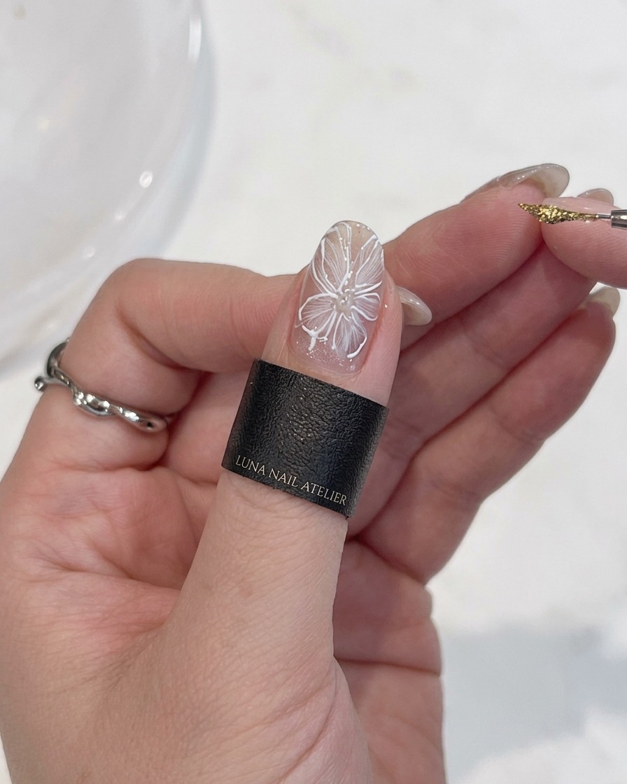 A close-up view of a hand holding a fine detail brush, applying a precise dot of metallic gold gel to the center of a painted white flower on a fingernail.