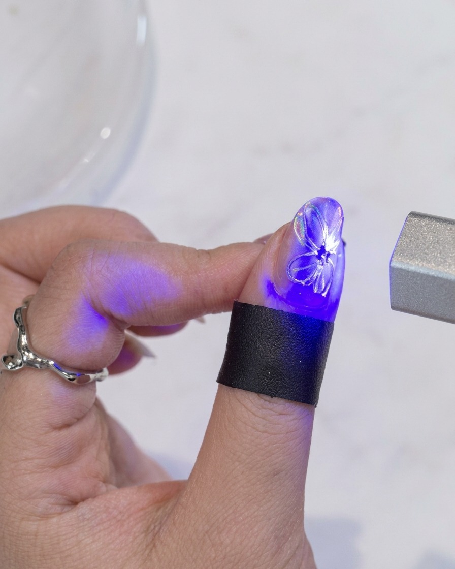 A brush applying a final layer of clear top coat over a nail featuring a white flower design with a gold center.