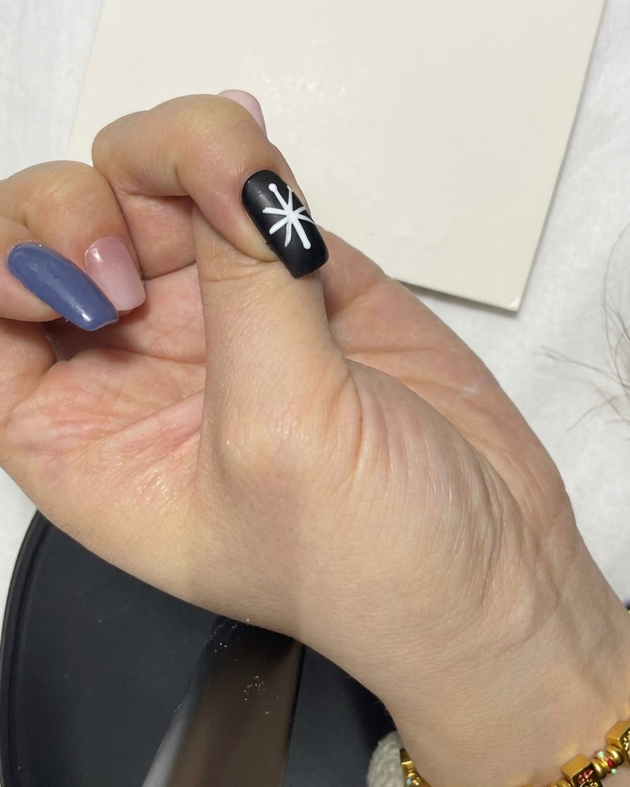 A hand using a liner brush to refine the white line segments forming a flower shape on a black nail.