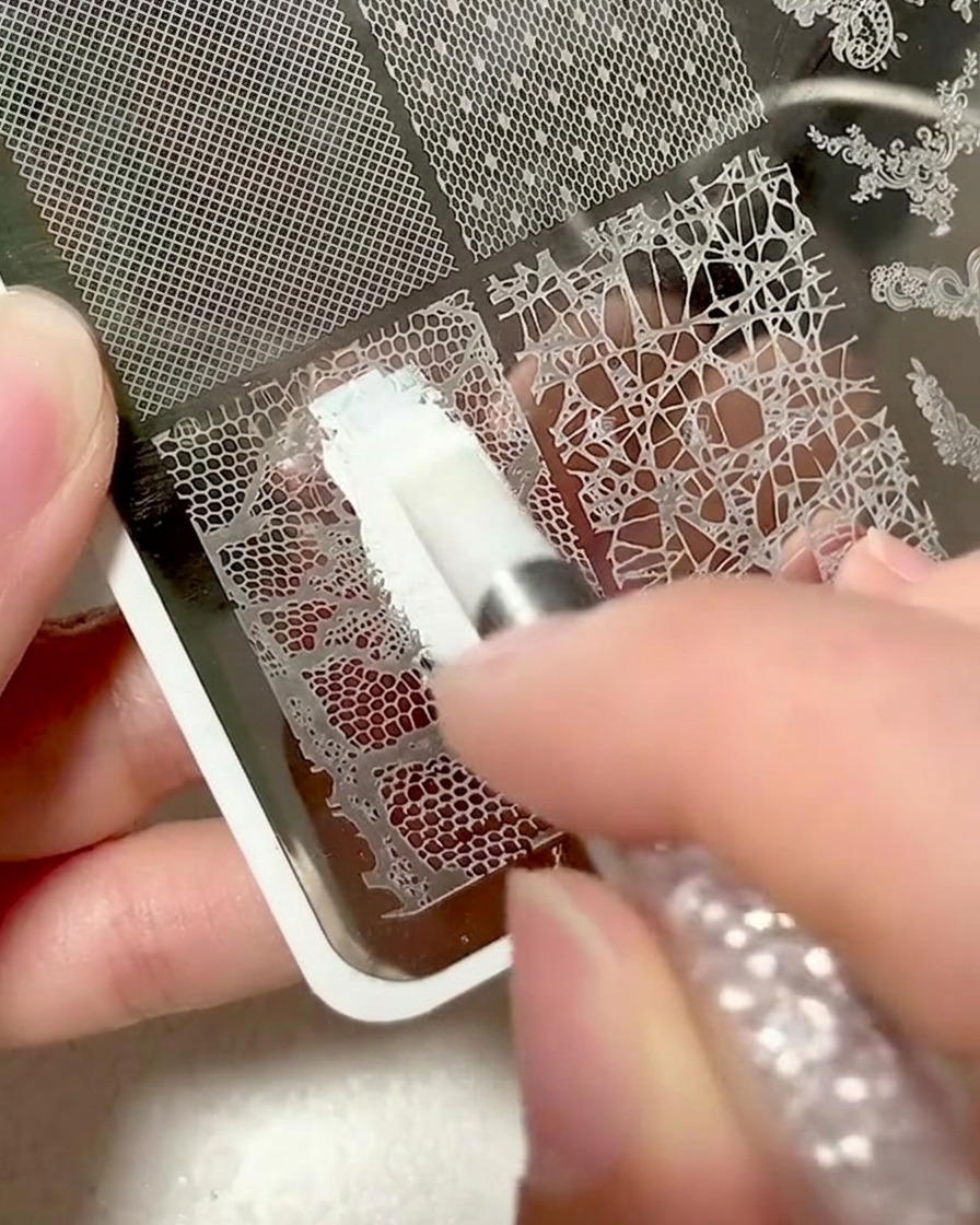 A close-up of a metal plate with a lace design being covered in white stamping gel.