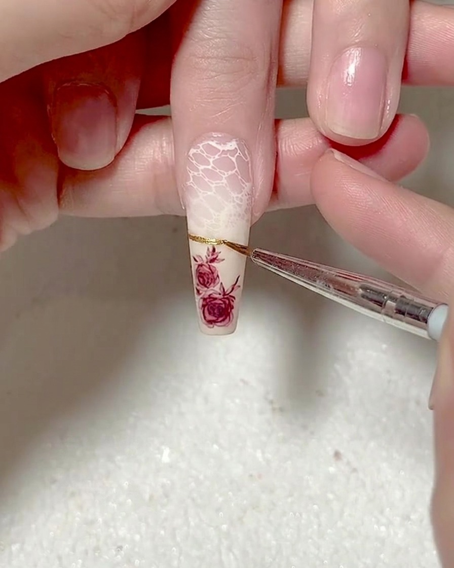 A close-up of a liner brush applying a thin, precise gold metallic gel French tip line across a nail.