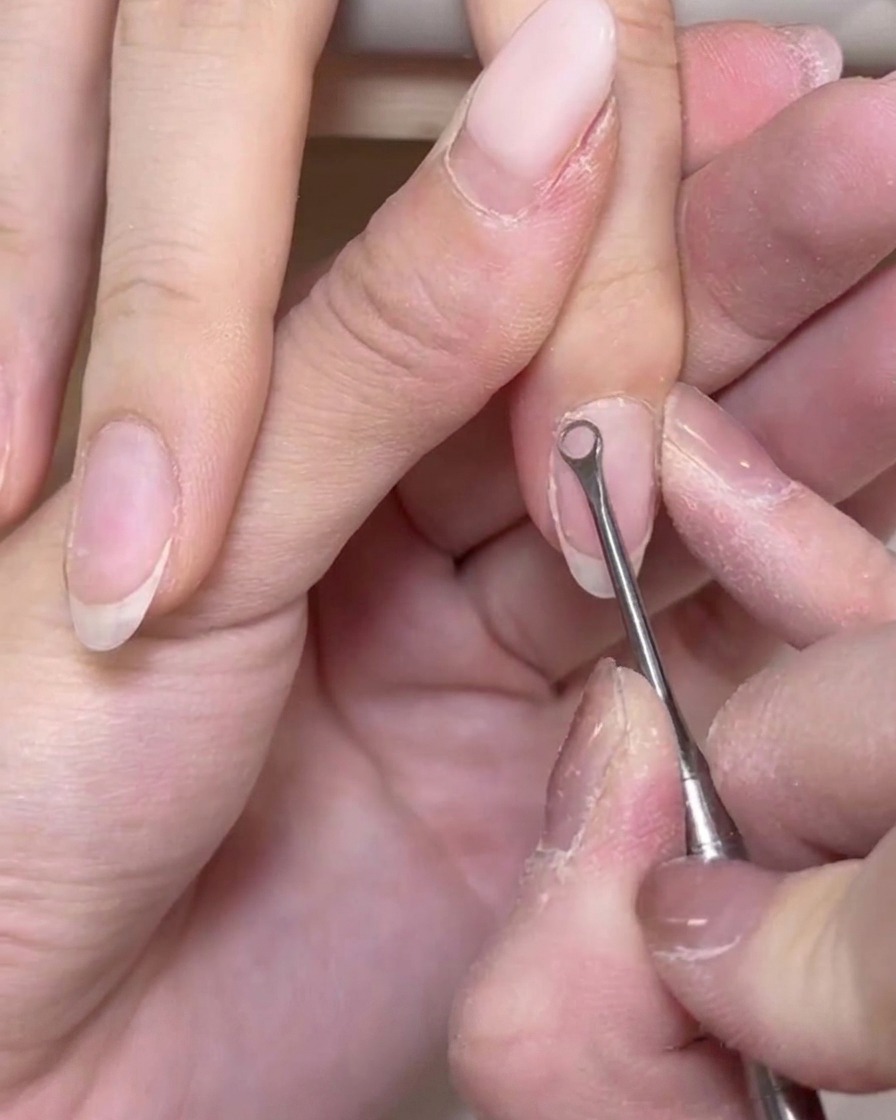 A close-up view of a metal tool gently scraping dead skin cells off the surface of a natural nail.