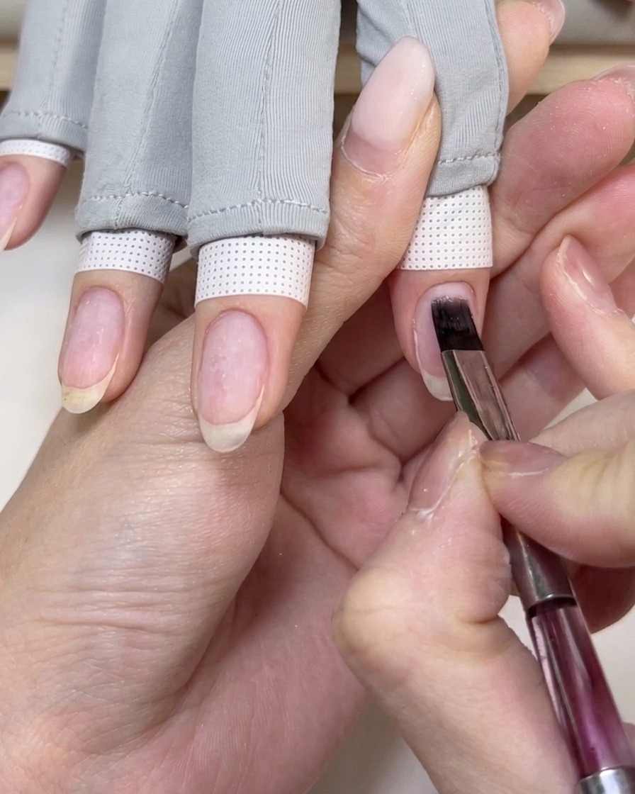 A flat nail gel brush applying a clear base coat onto a natural fingernail.