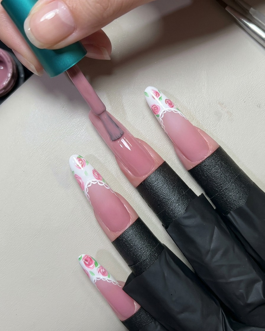 A polish brush applying a coat of translucent nude pink gel over a long prepared fingernail.cite: 1