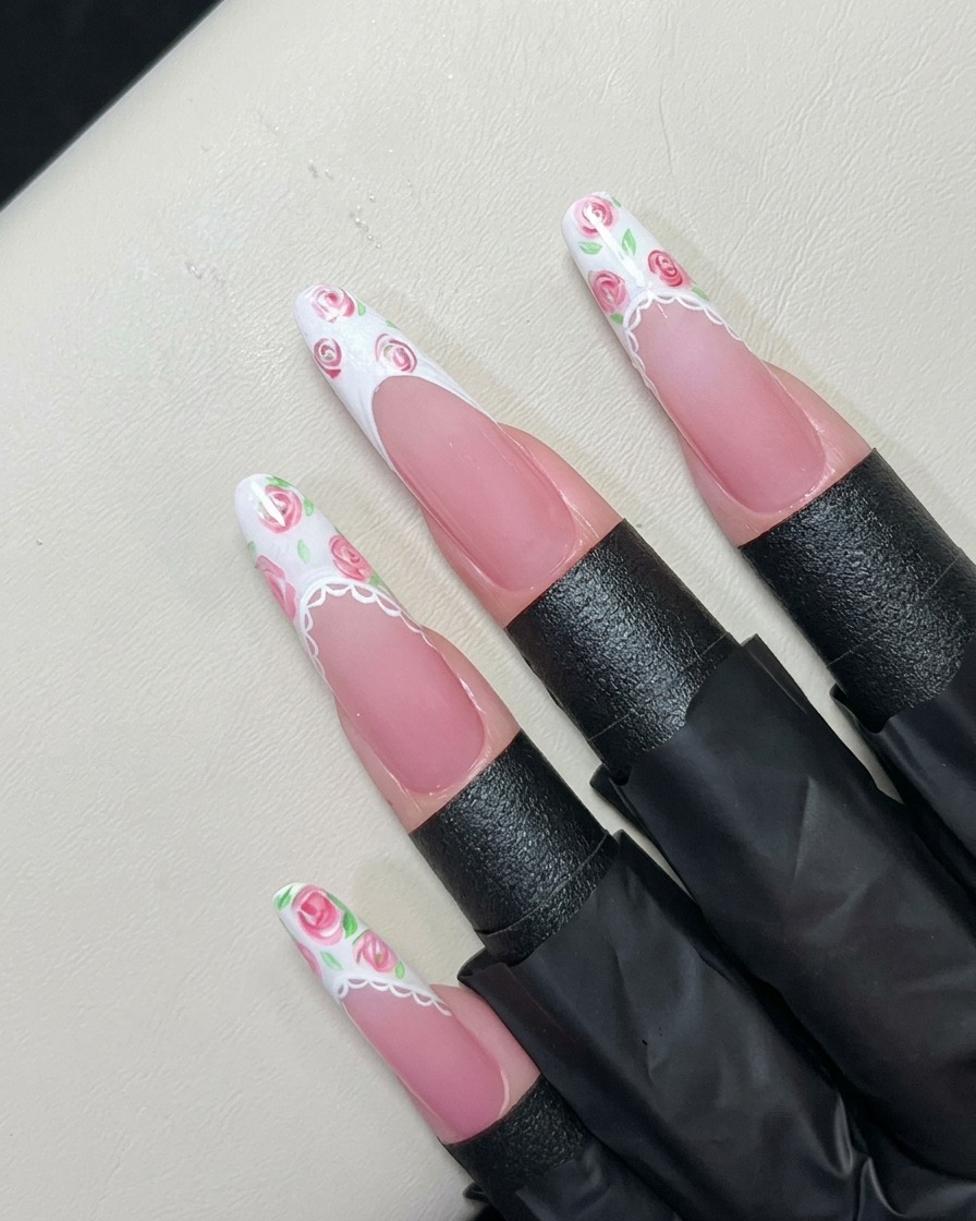 A fine liner brush painting tiny green leaves around pink roses on a white gel French tip.cite: 1