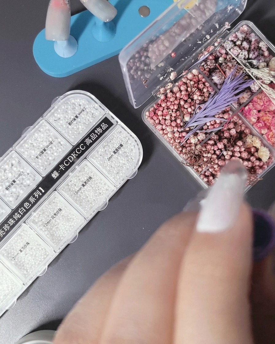 Tweezers holding a small dried flower petal, placing it onto a nail surface with wet gel.