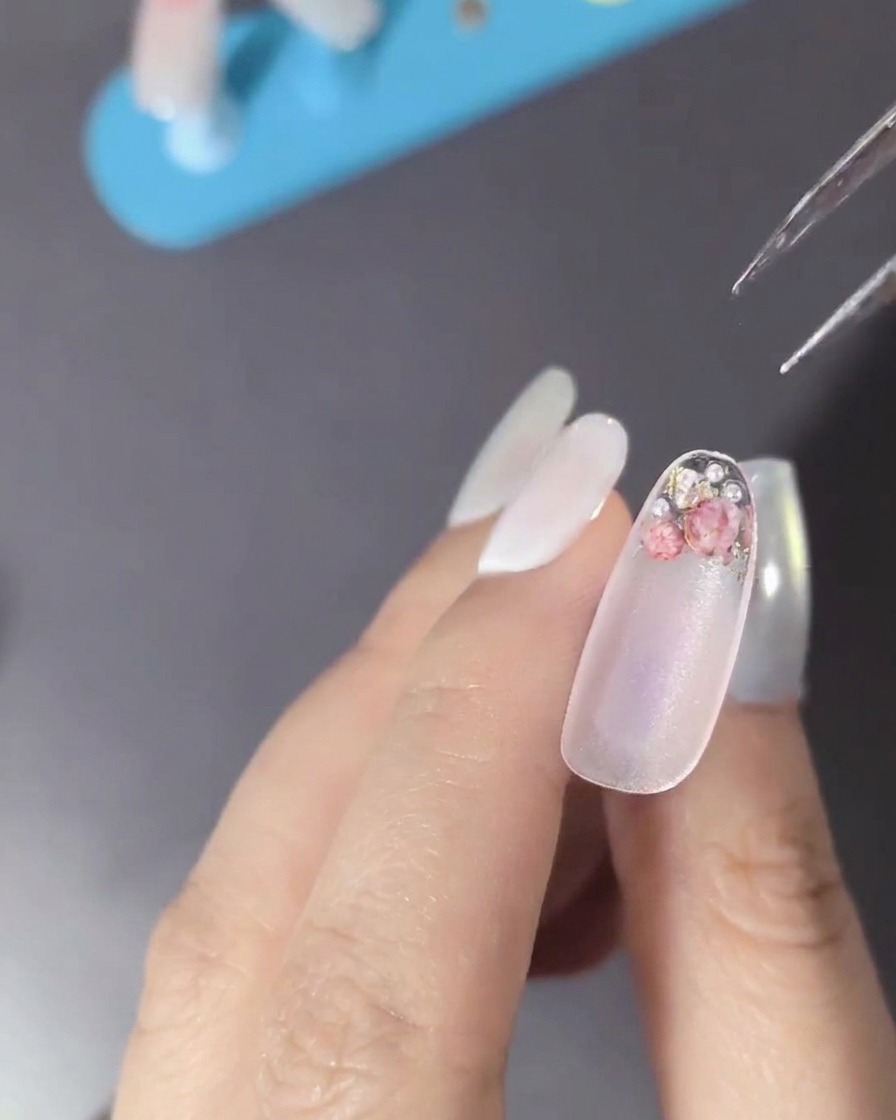 A close-up of a nail decorated with dried flowers, pearls, and small metallic rhinestones.