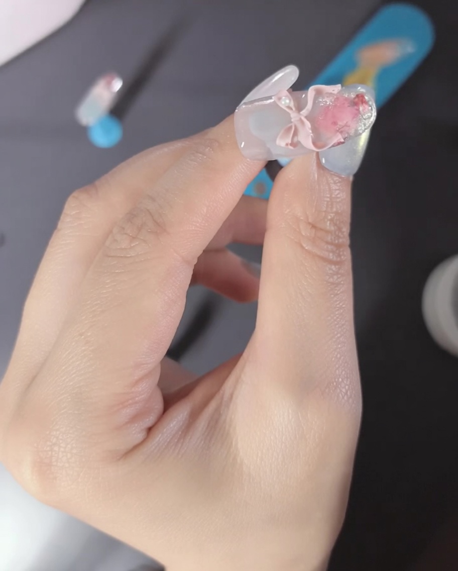 A close-up of a finger featuring a pink 3D ribbon bow charm and pearl accents on the nail.