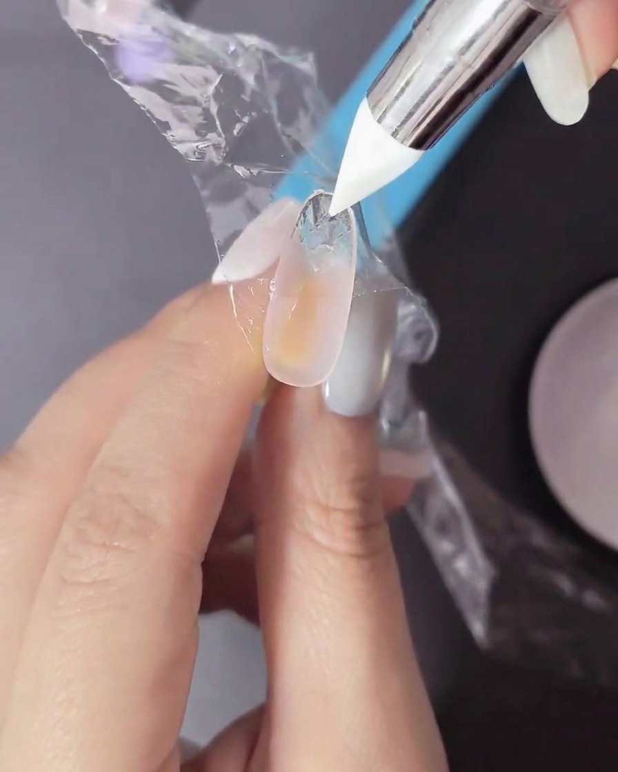 A finger with an uncured gel application has a piece of thin plastic film being pressed onto the nail surface by a silicone tool.