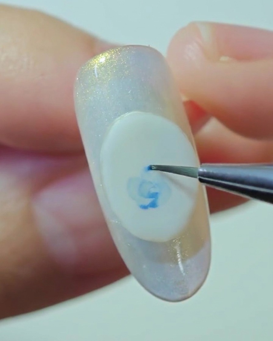 A close-up view of a detail brush gently blending the edges of a blue rose painted on a white gel base.