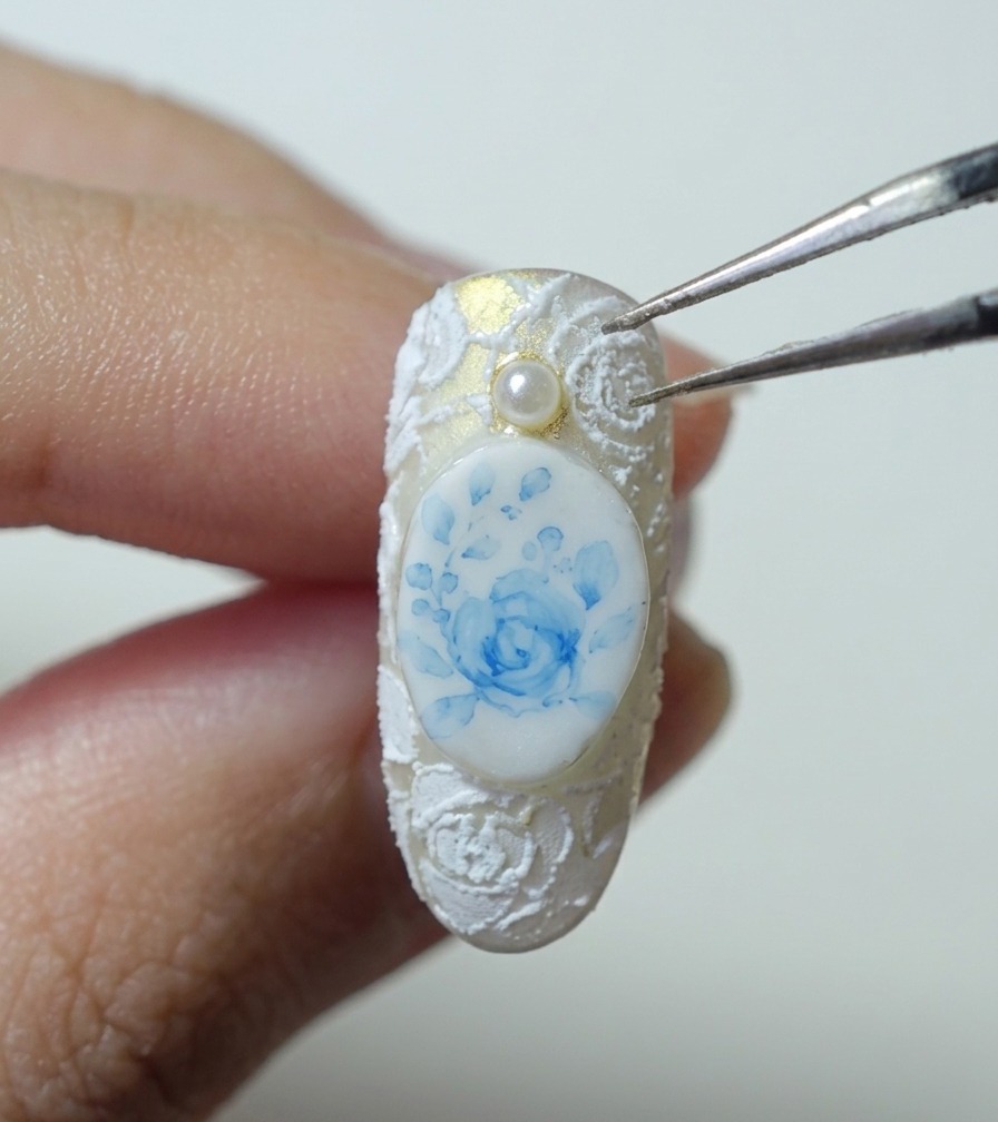 Tweezers holding a small white pearl bead above a nail with blue rose and white lace relief art.