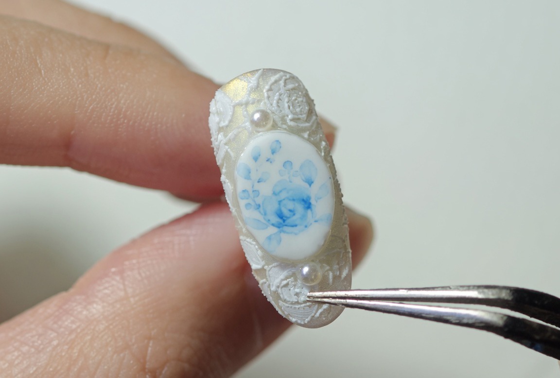 A close-up of a decorated nail with blue roses and white lace, being held by tweezers, ready for final curing.
