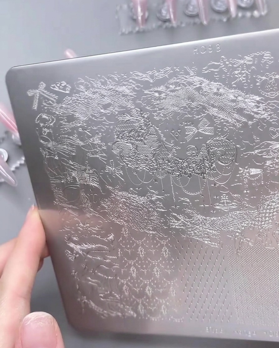 A close-up view of a metal nail stamping plate with a white gel polish being spread over a detailed lace pattern using a scraper.