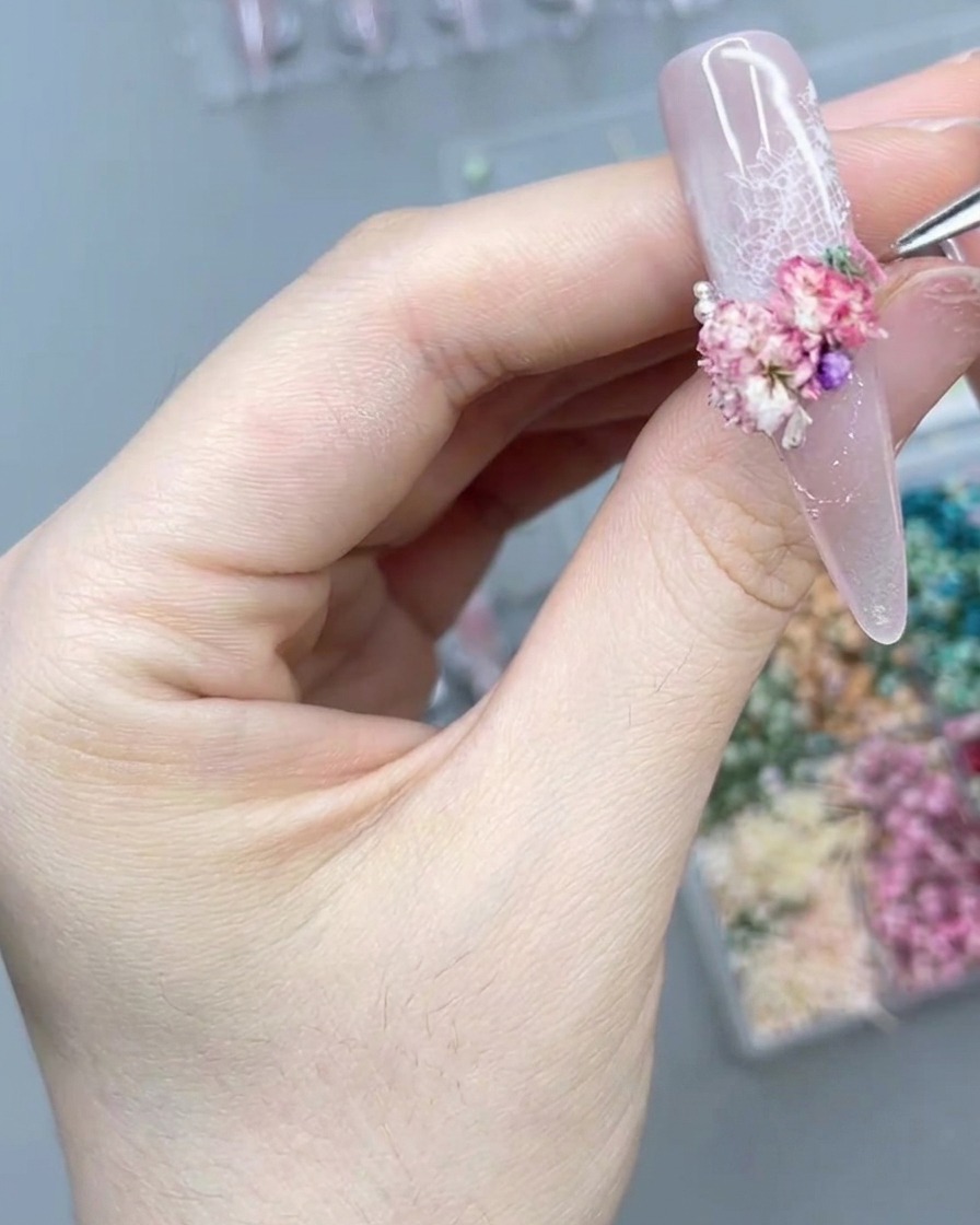 A close-up of a hand using a precision tool to carefully place a tiny pearl next to a cluster of pink dried flowers on a long press-on nail.