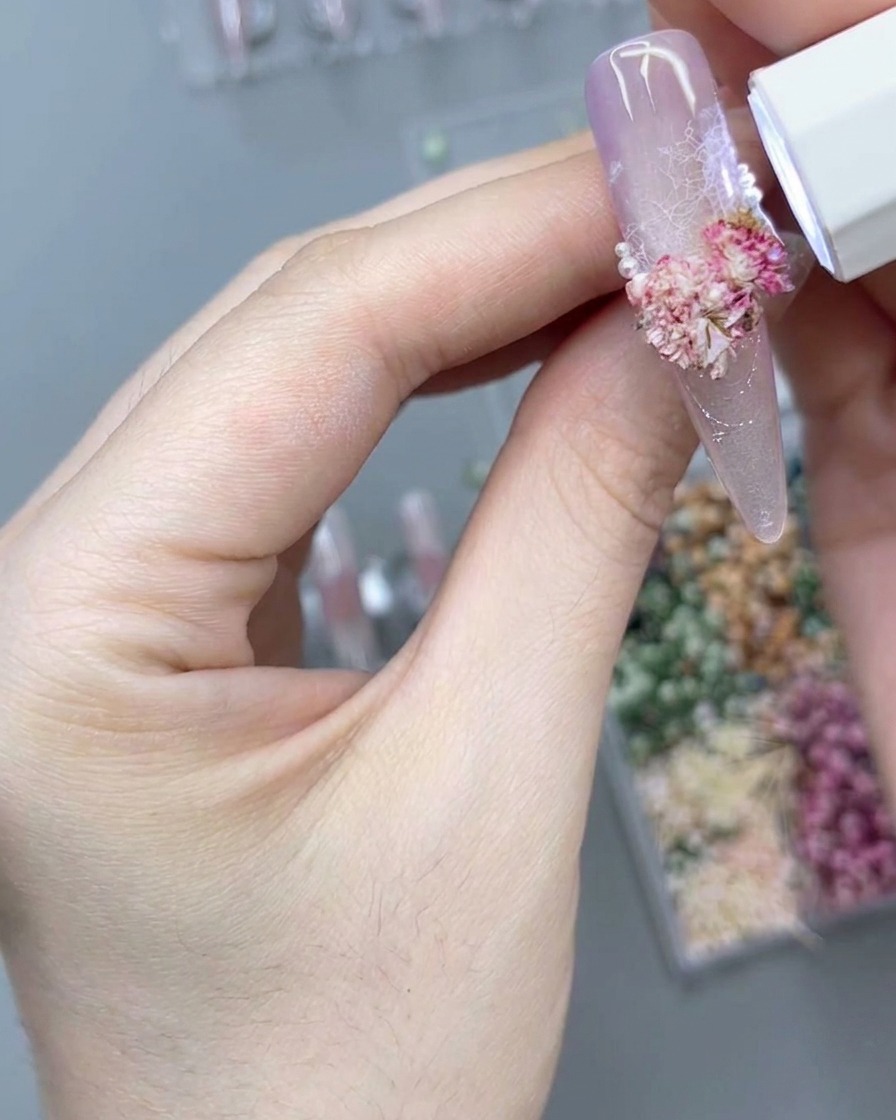A portable white UV/LED lamp shining downward over a 3D pink floral arrangement on a long pink press-on nail to cure the design.