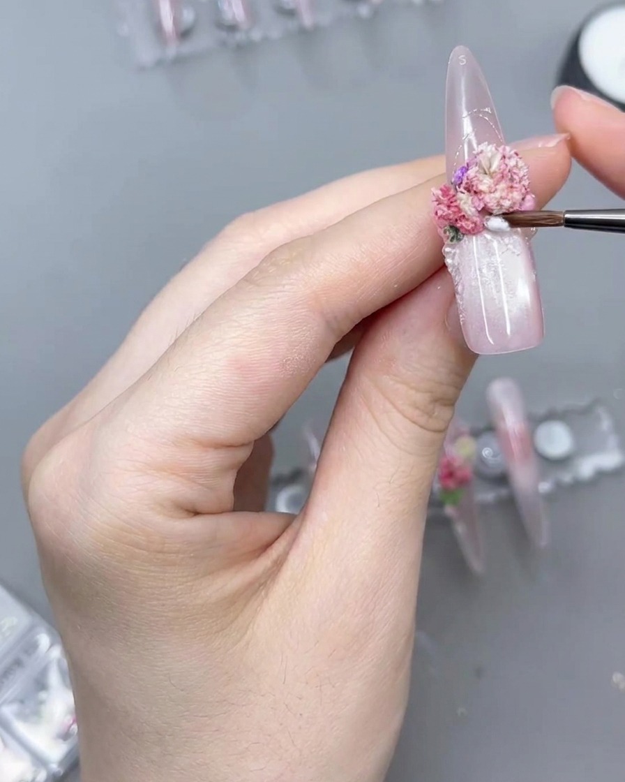 A close-up of a fine detail brush applying clear product around the base of a pink floral cluster on a long press-on nail.