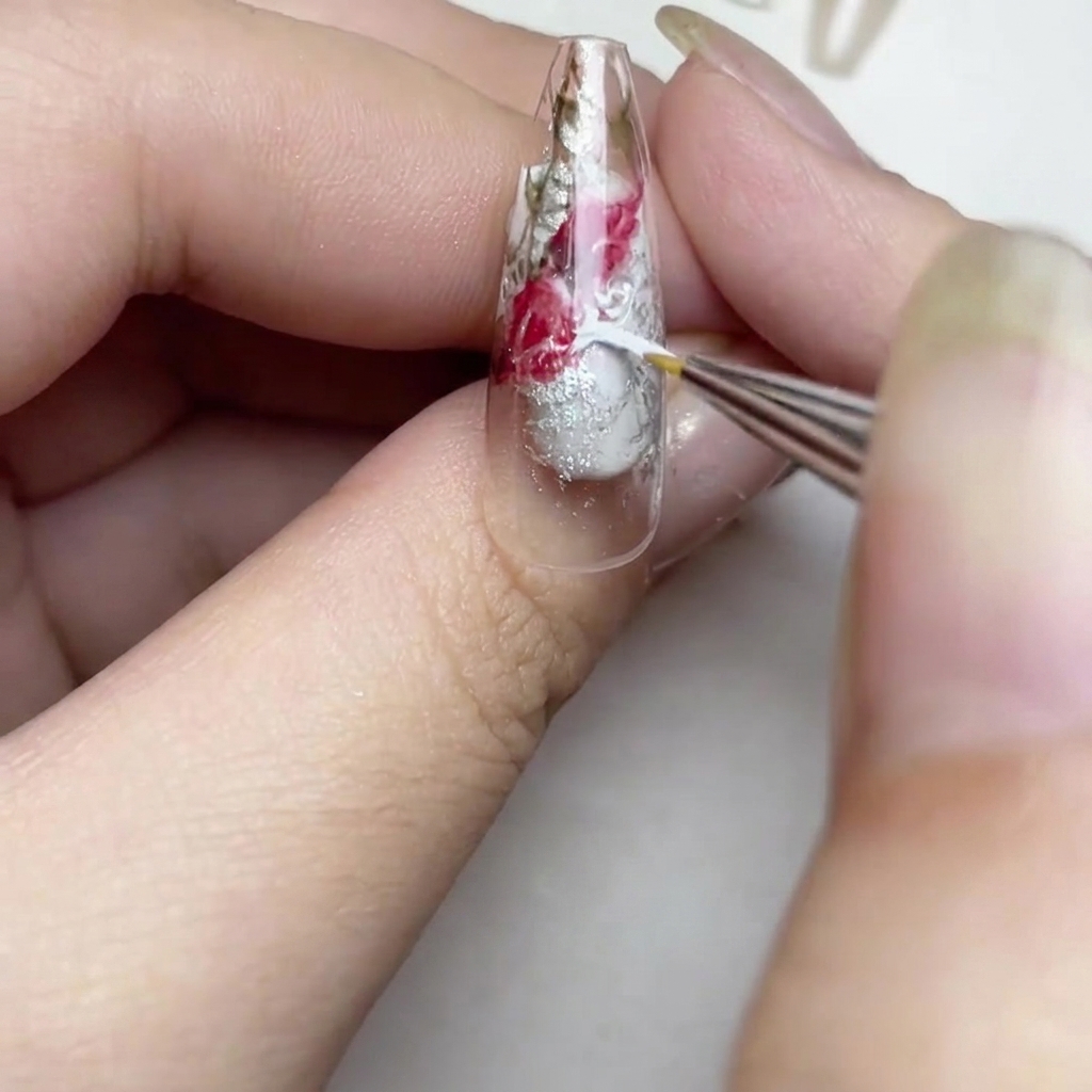 A close up of a nail showing a brush adding thin white paint strokes to create a soft watercolor rose effect in the clear spaces.