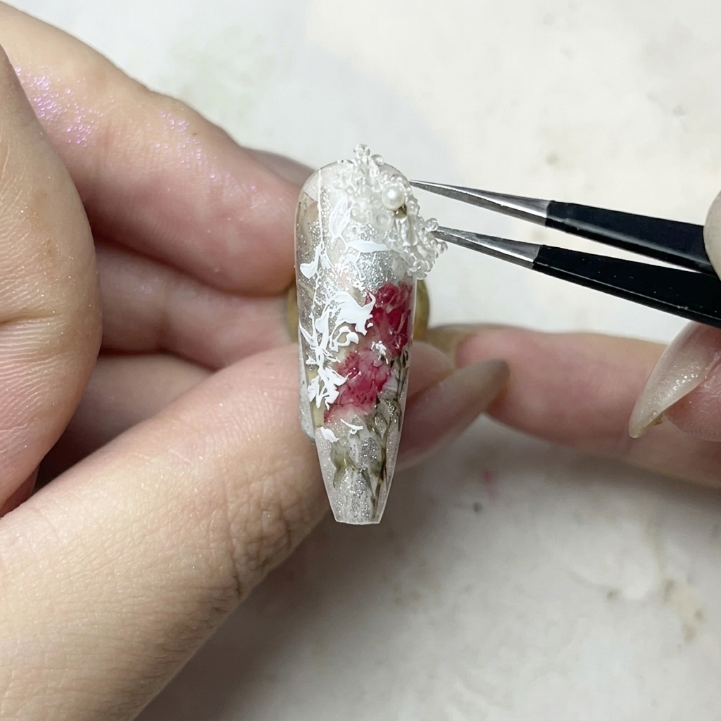 A pair of tweezers carefully placing a small, white 3D flower charm onto a dot of clear gel glue on a manicured nail.