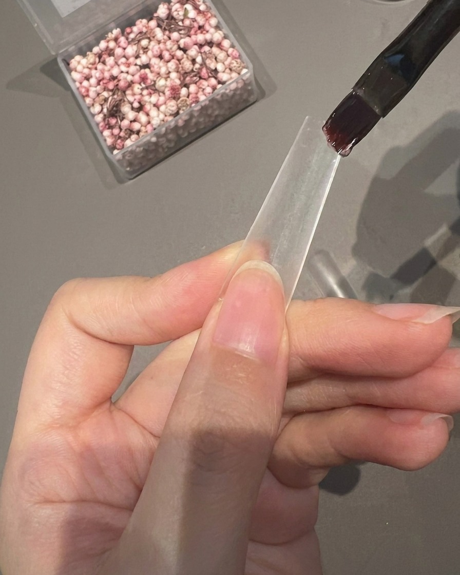 A brush applying a layer of clear adhesive gel onto the bottom half of a clear nail tip.