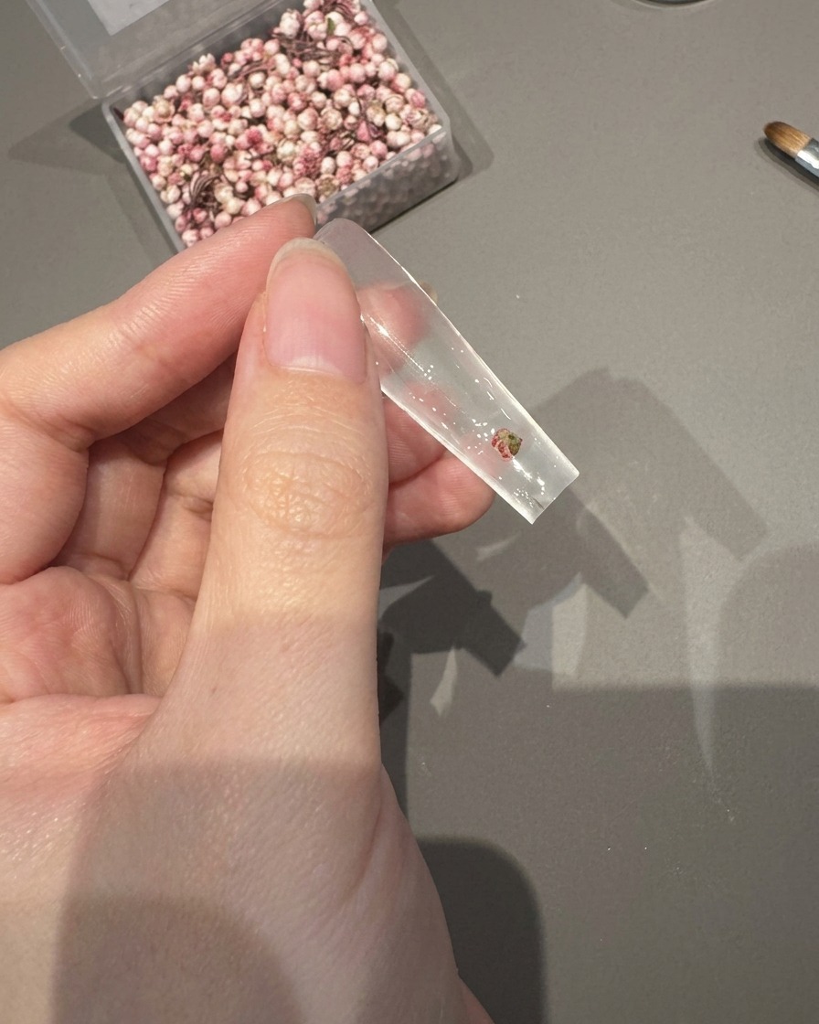 Tweezers placing a small dried flower onto a clear nail tip coated in wet adhesive gel.