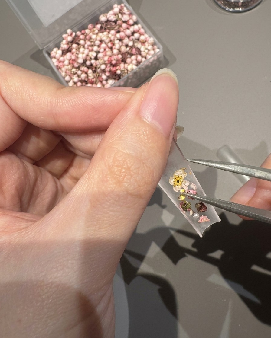 Close-up of tweezers placing tiny pearls and crystals onto a nail tip decorated with dried flowers.