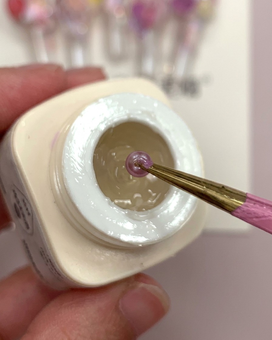 A hand holding a clear nail tip with a drop of frosted glue and purple flower balls being placed into it.