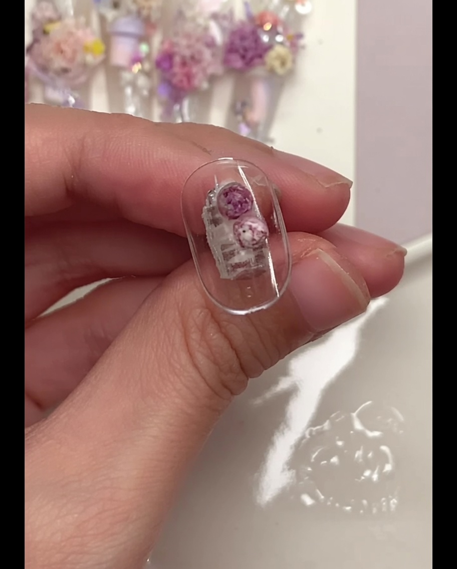 A detail brush gently pressing down on purple flower balls placed on a clear nail tip.