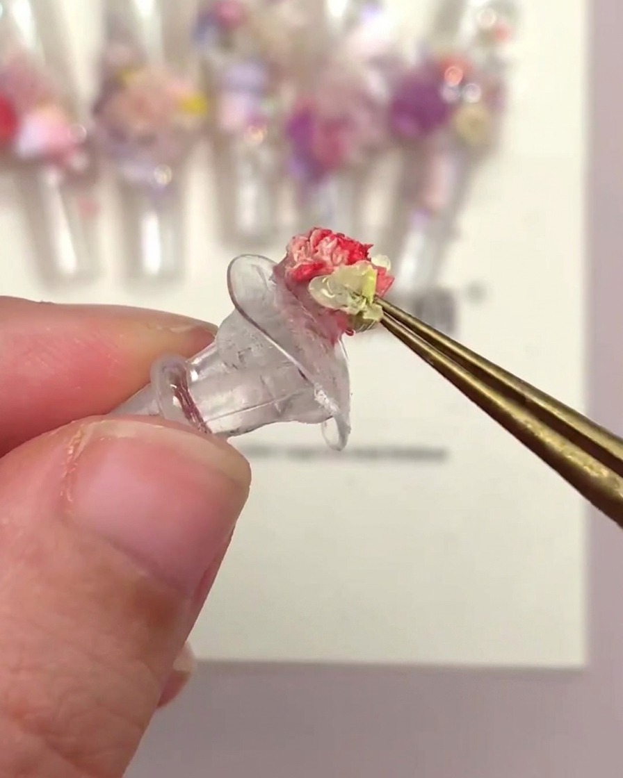 A hand using tweezers to place a trumpet-shaped flower head onto a nail tip decorated with other flowers.