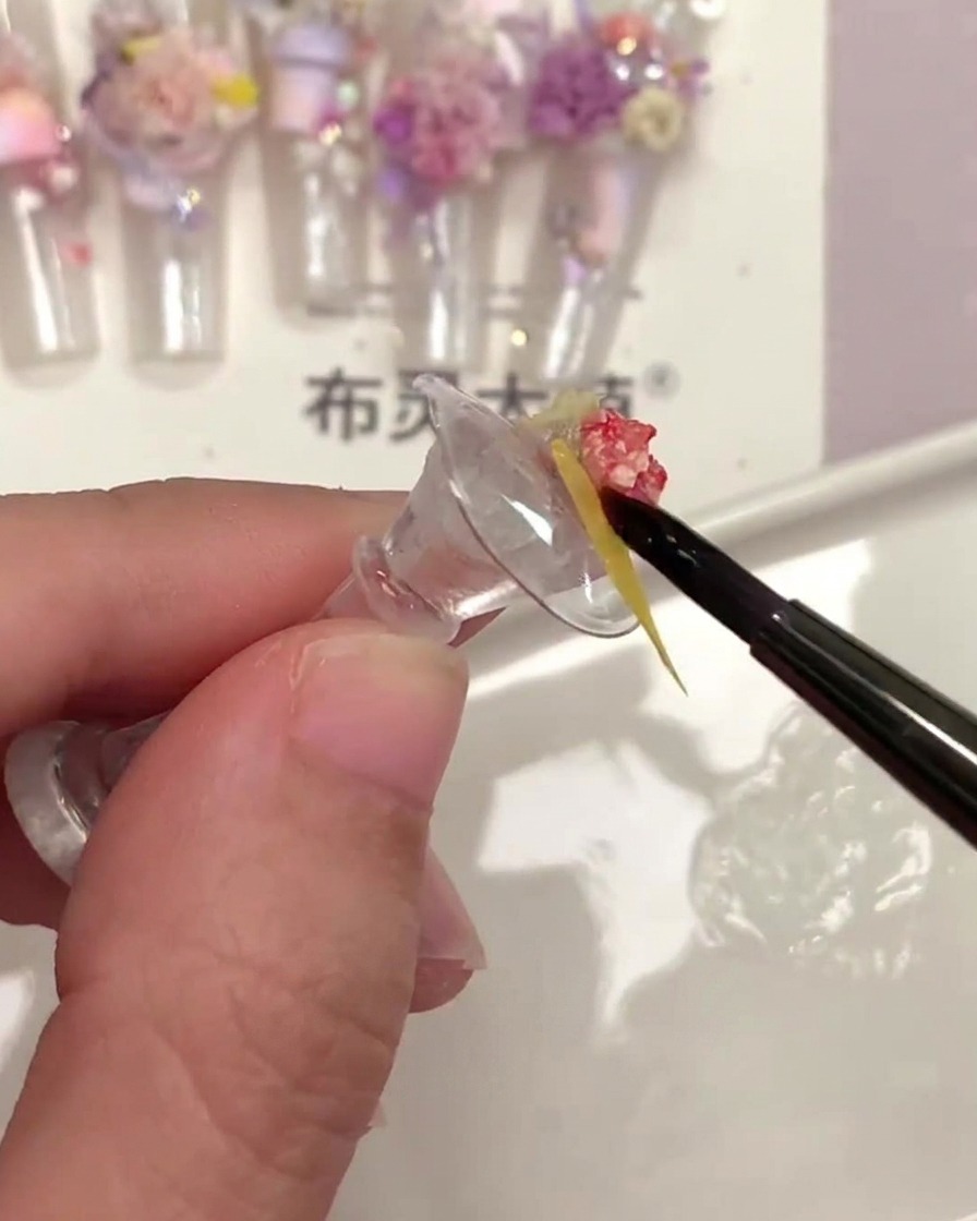 A fine nail brush applying clear top coat underneath a small yellow dried flower piece.