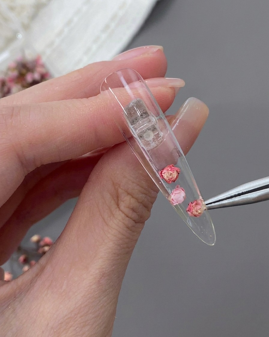 Fine point tweezers placing a small pink dried flower next to a larger central rose inside the back of a clear stiletto nail tip.