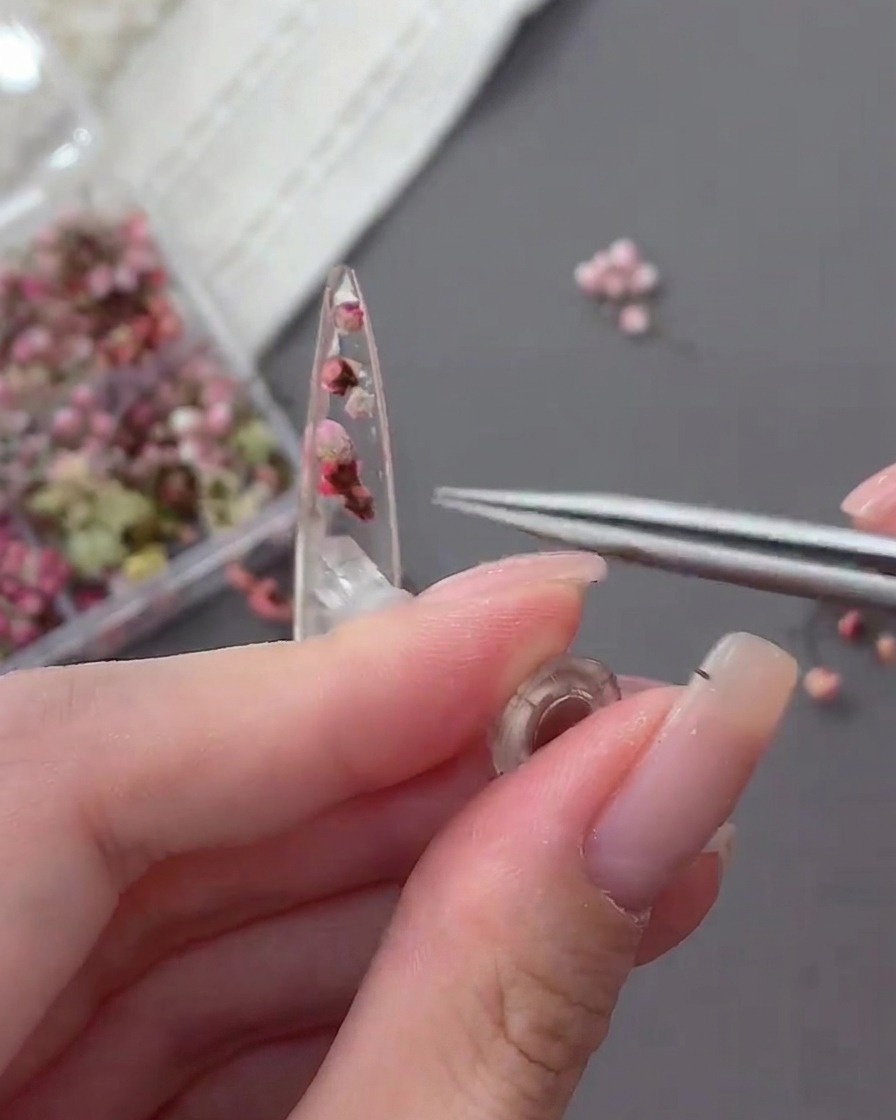 A close-up of a pair of tweezers placing a tiny clear rhinestone onto the surface of an uncured nail tip containing dried flowers.
