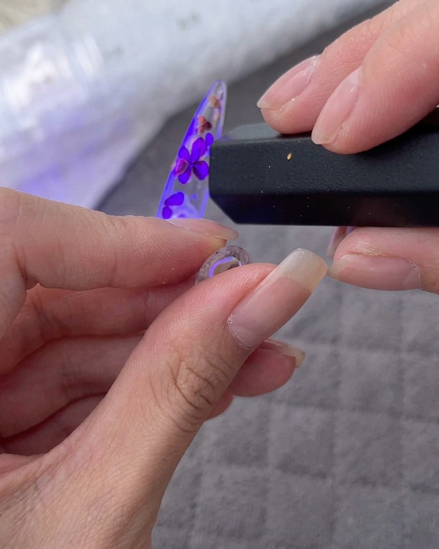A hand holding a handheld UV/LED lamp over a nail tip decorated with flowers and rhinestones.
